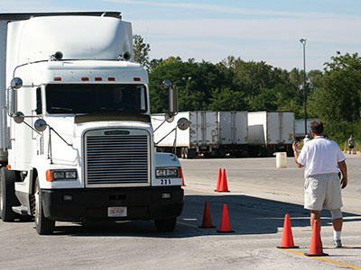 image off semi-truck navigating a road course with orange cones with a man off to the side giving directional hand signals